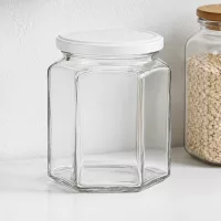 Leeres 400g Honigglas sechseckig mit weißem Deckel neben Vorratsglas mit Haferflocken auf weißer Küchenablage