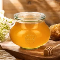 WECK Schmuckglas 560ml RR80 mit Honig befüllt neben Honiglöffel Bienenwaben und weißen Blüten auf Holzbrett