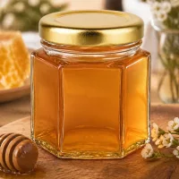 60g Honigglas gefüllt mit goldenem Honig neben Holzlöffel und Blüten auf rustikalem Holztisch dekoriert