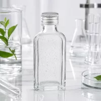 Miniatur Meplat Glasflasche 50ml klar mit silbernem PP22 Aludeckel neben Laborgeräten